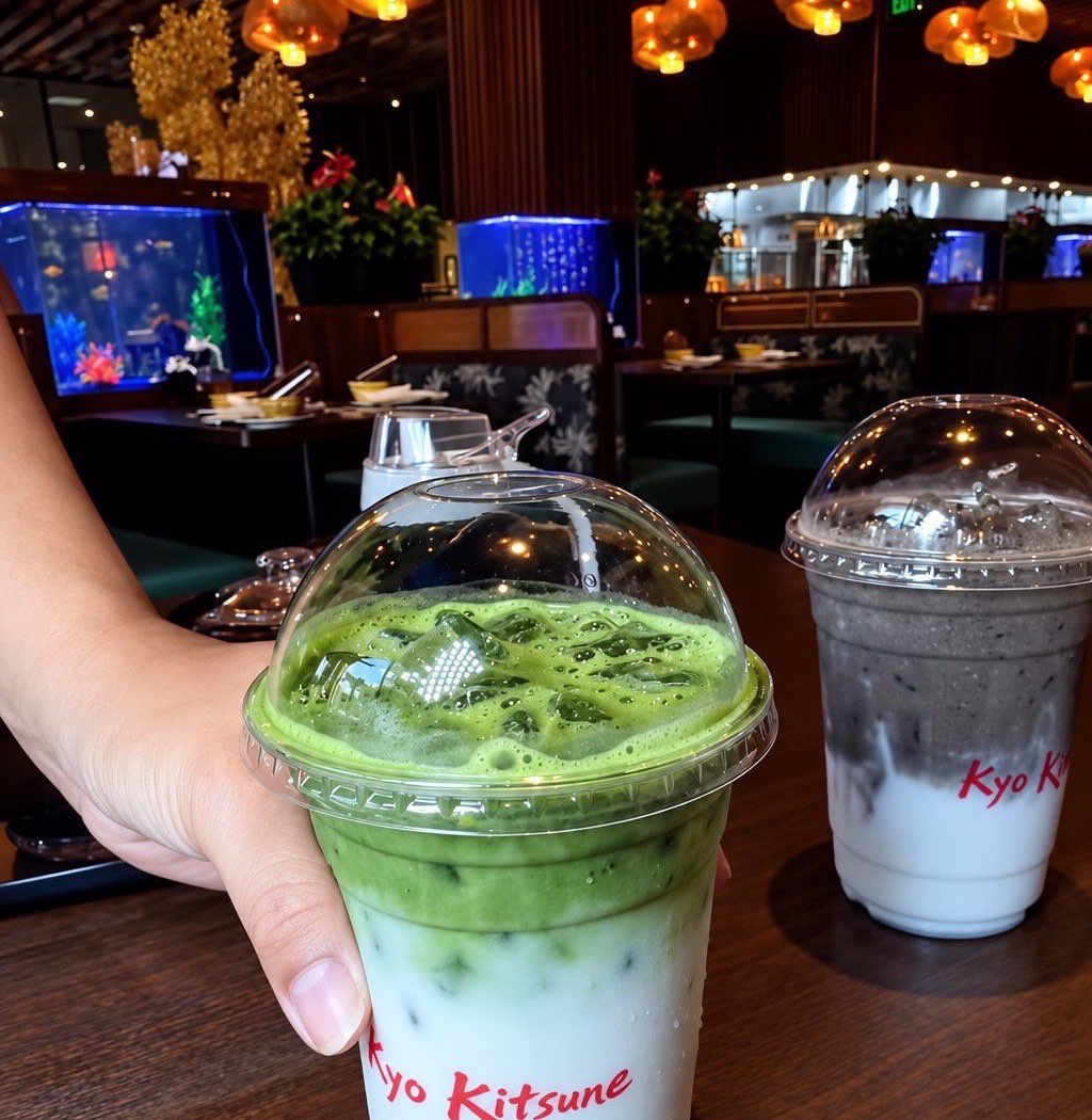 Iced matcha latte in the tea room A hand holding a tall iced matcha latte in a printed Kyo Kitsune cup inside the tea room, warm lantern light overhead