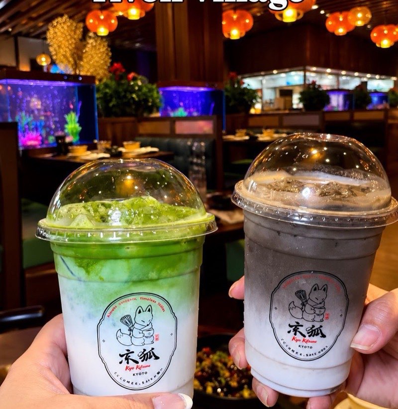 Guests toasting with Kyo Kitsune drinks Two guests raising Kyo Kitsune cups together, one iced matcha and one dark sesame drink, both printed with the fox emblem